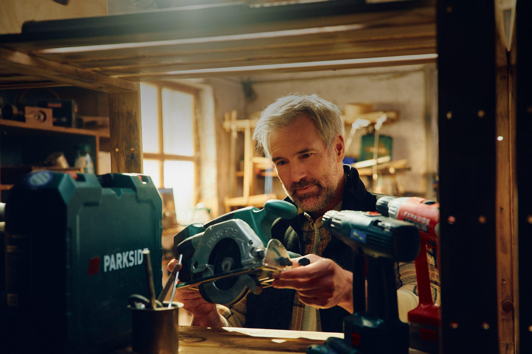 A man examines a circular saw in a workshop, with power tools nearby.
