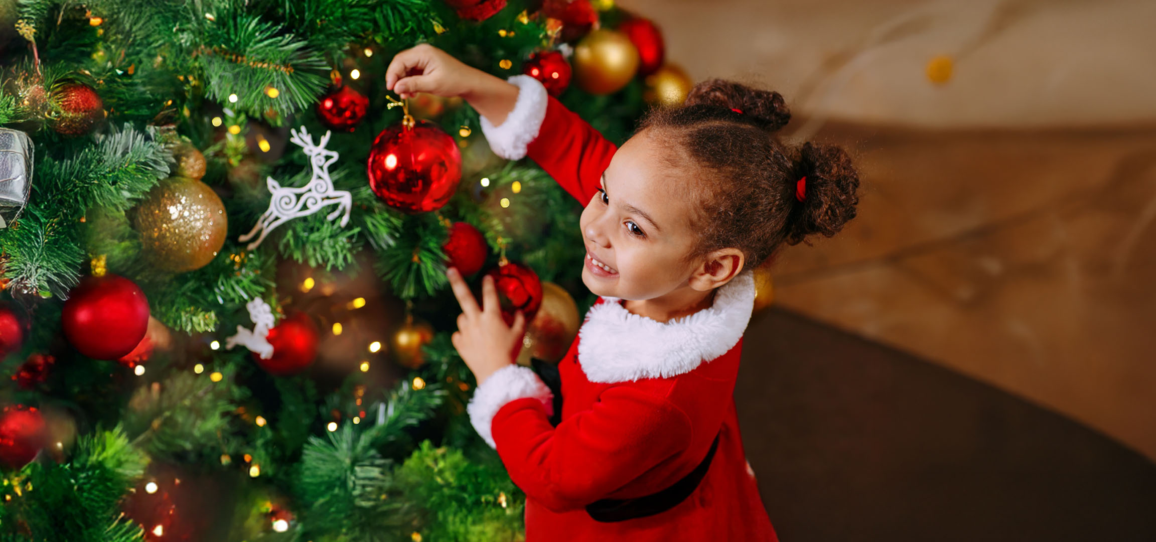 Happy little girl in Santa costume decorating a Christmas tree with red baubles.