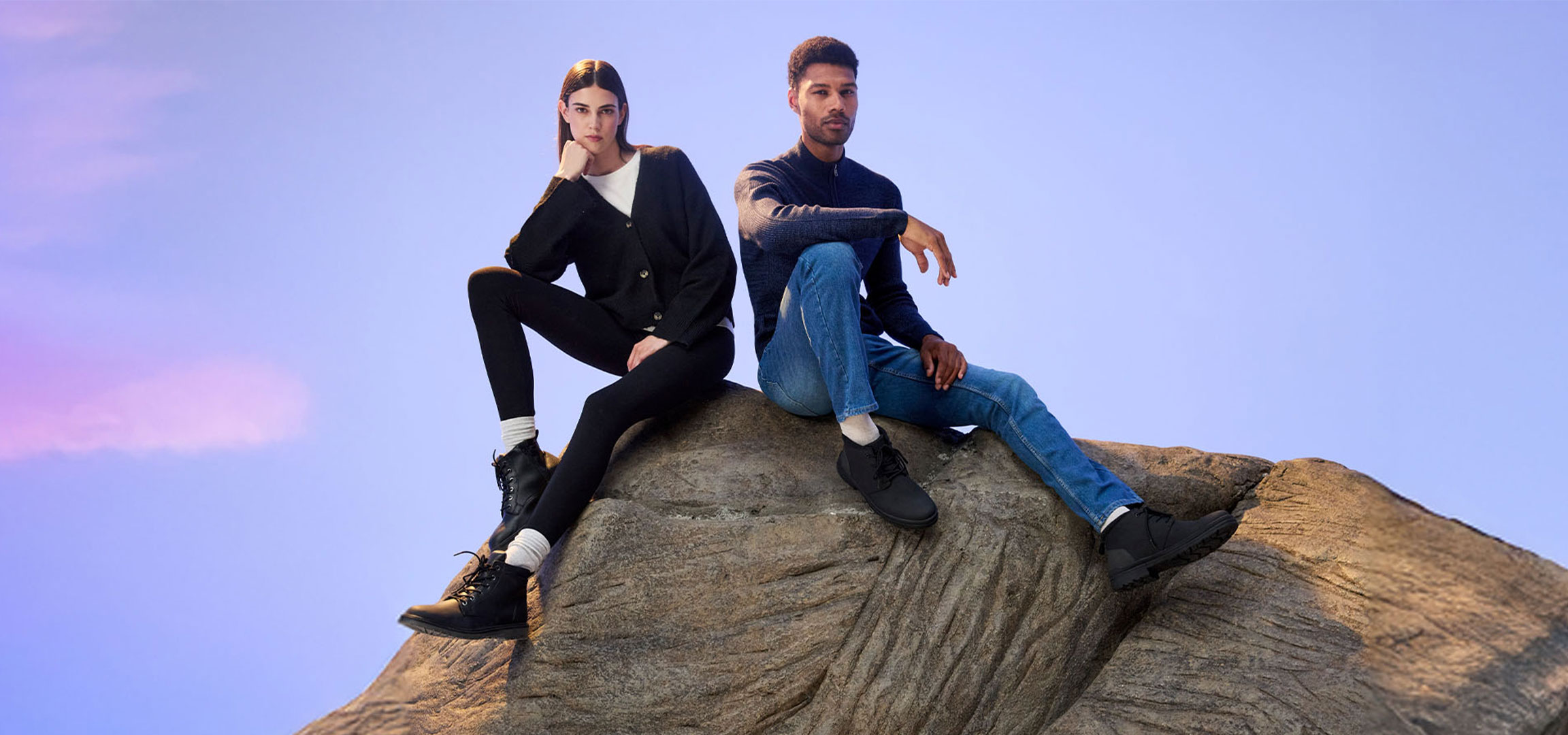 Two people in casual clothing sitting on a rock with a purple sky.