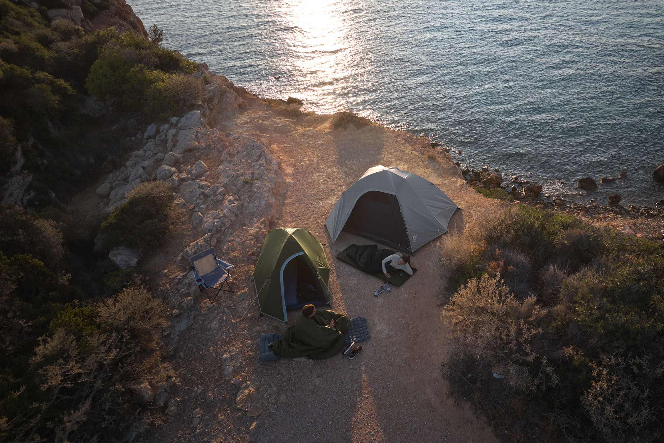 Two people sleeping in sleeping bags next to camping tents on a rocky coast at sunset.