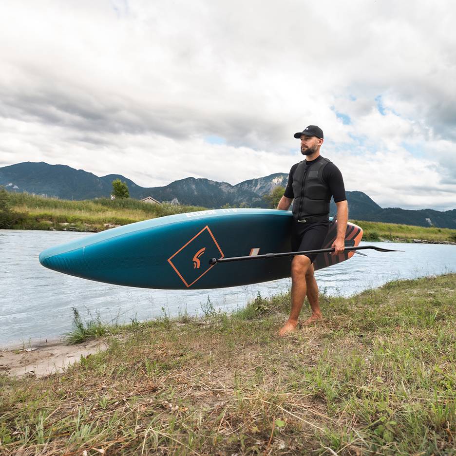 Man in a life vest and cap, carrying a paddleboard and paddle by a river.