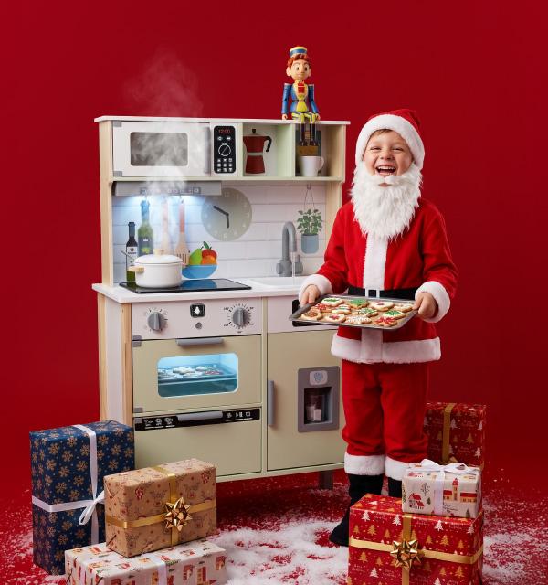 A child in a Santa suit holding a tray of cookies next to a wooden toy kitchen and gifts.