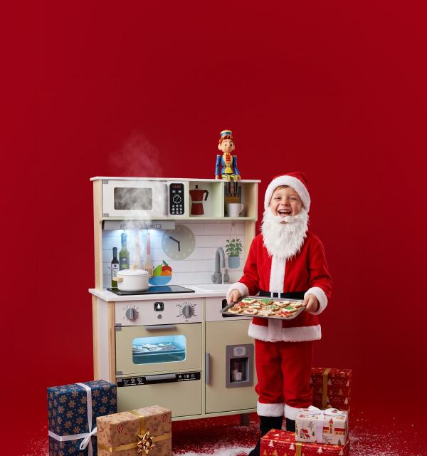 A child dressed as Santa Claus holding a tray of cookies next to a wooden toy kitchen.
