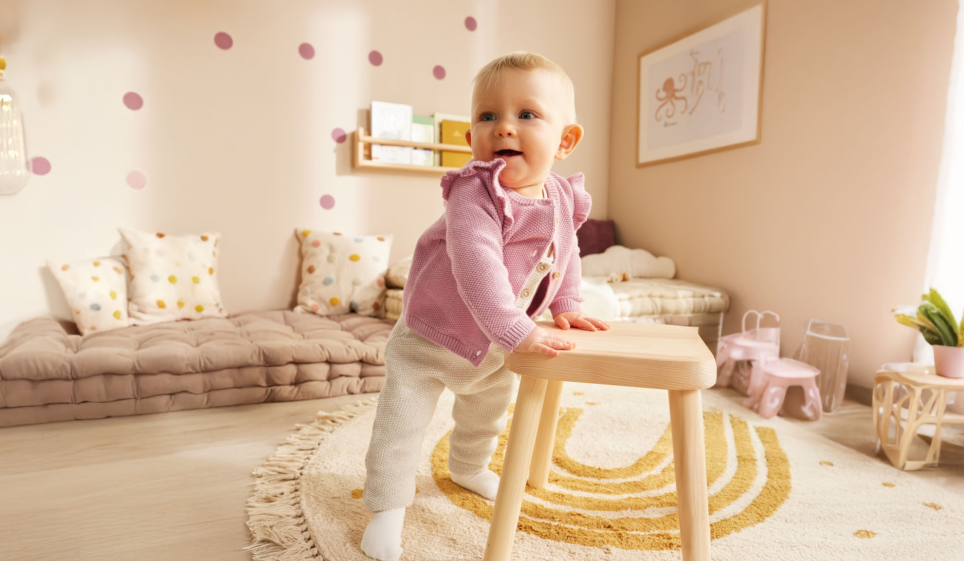 A toddler stands by a wooden stool in a minimally decorated nursery.