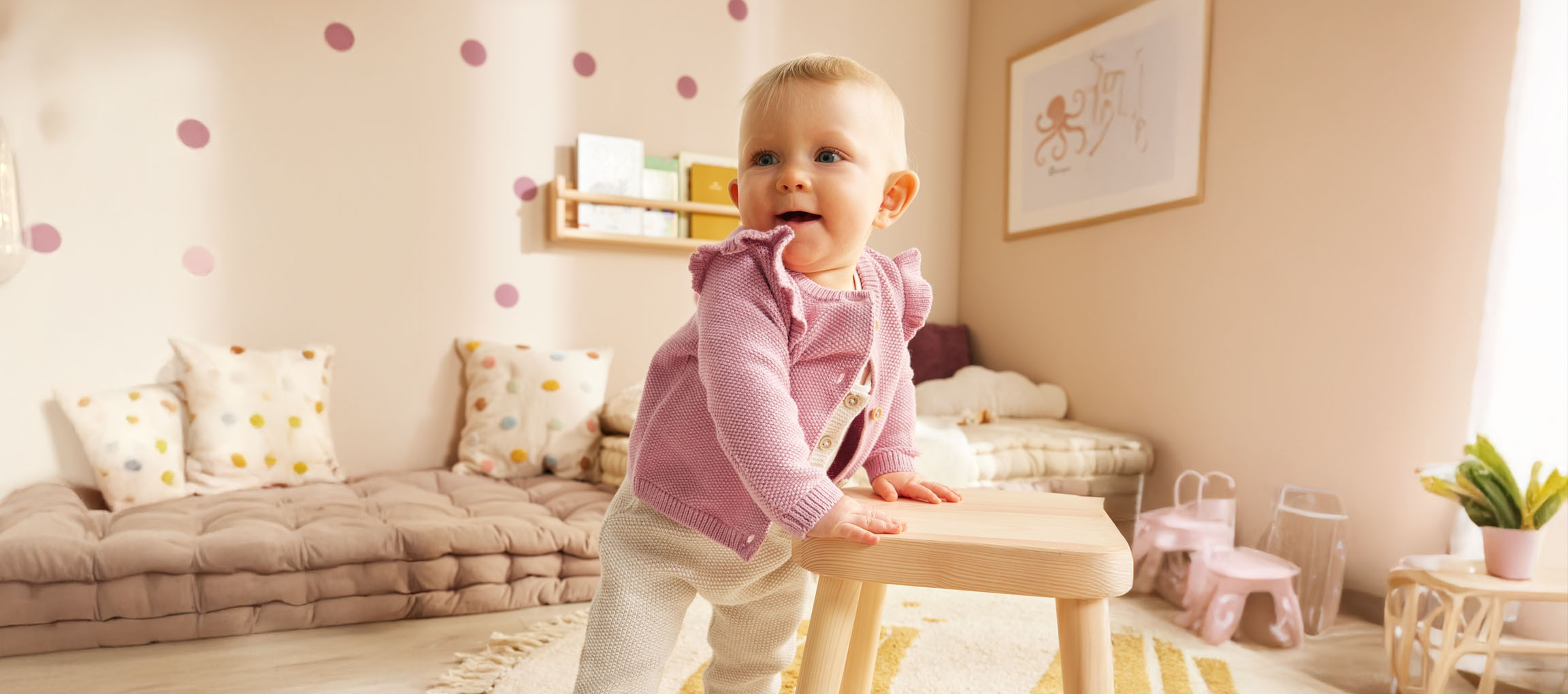 A baby in a cute pink cardigan stands near a wooden stool in their nursery.