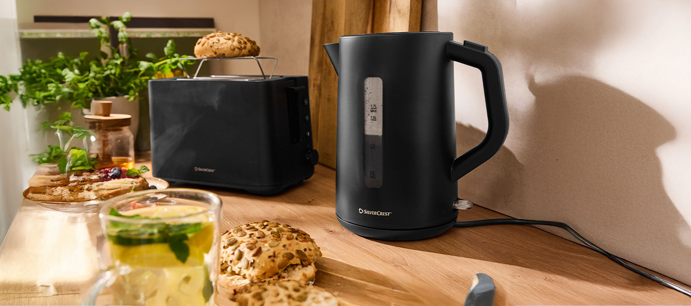 Black SilverCrest kettle and toaster on a wooden counter with bread rolls and drinks.