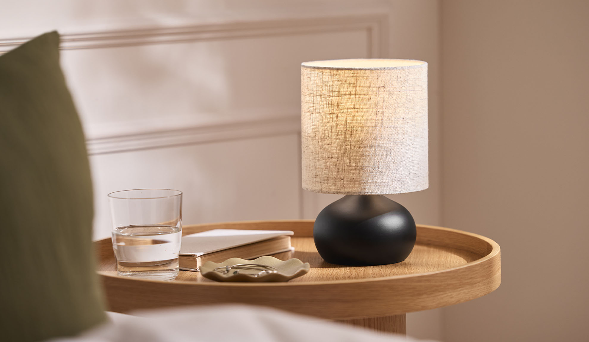 Bedside lamp, glass, book, and jewelry tray on a wooden tray next to the bed.