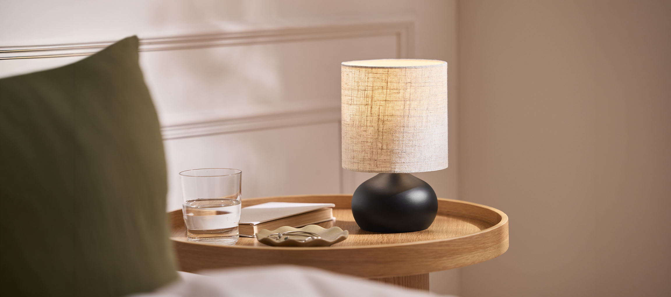 Bedside lamp with fabric shade, glass of water, and book on a wooden tray.