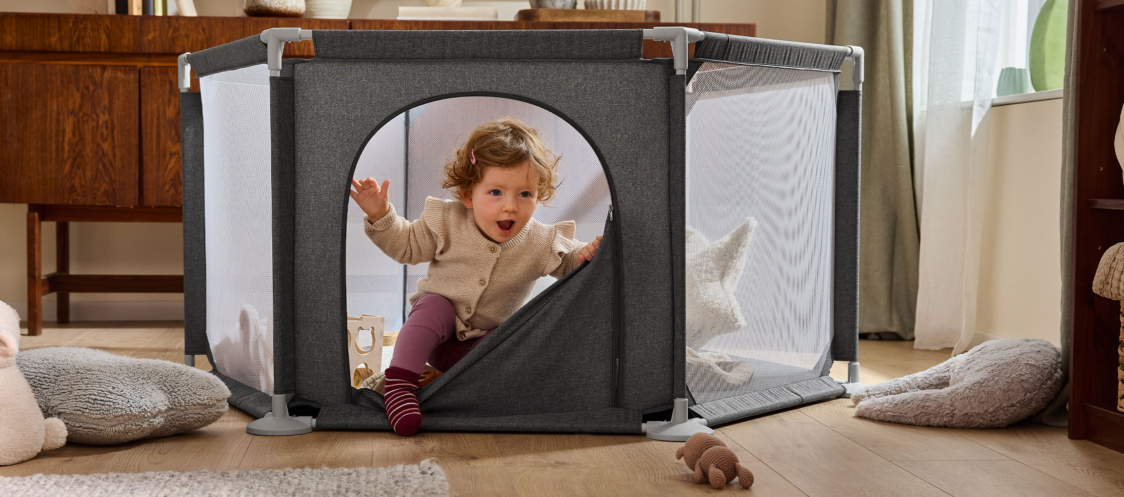Happy baby crawling out of a grey fabric playpen with mesh sides on a wooden floor.