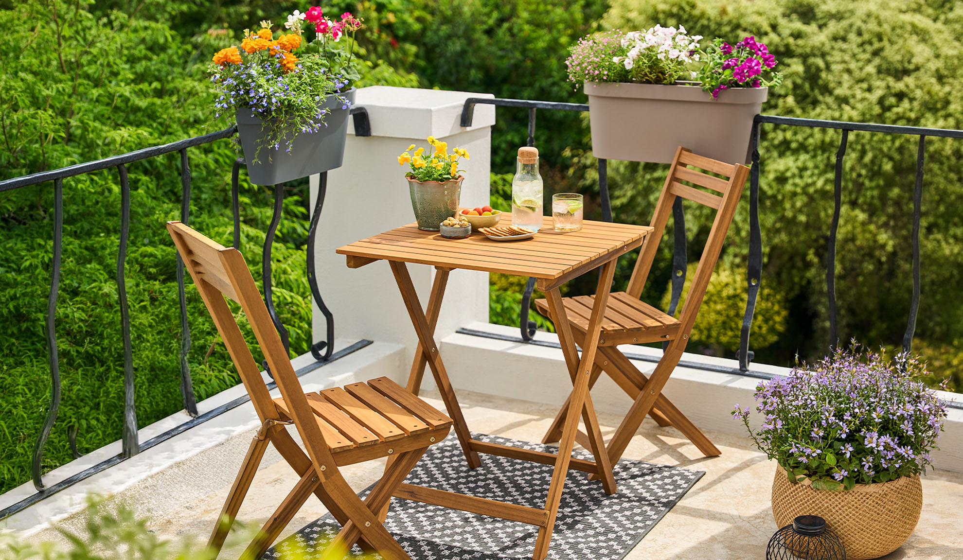 Balcony with wooden bistro set, potted flowers, and drinks on a patterned rug.