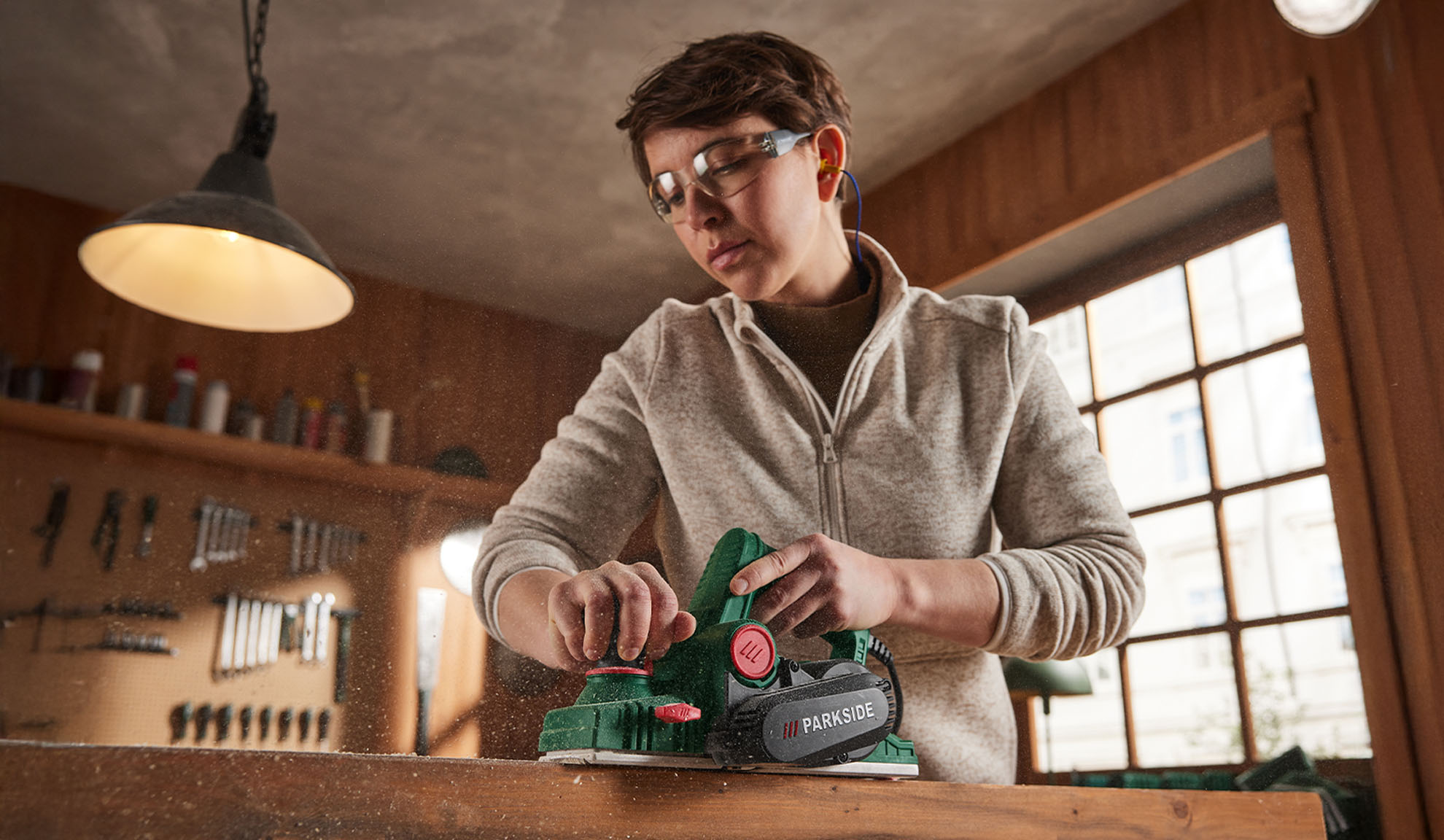 Woman with safety glasses and earplugs using a Parkside electric planer on wood.