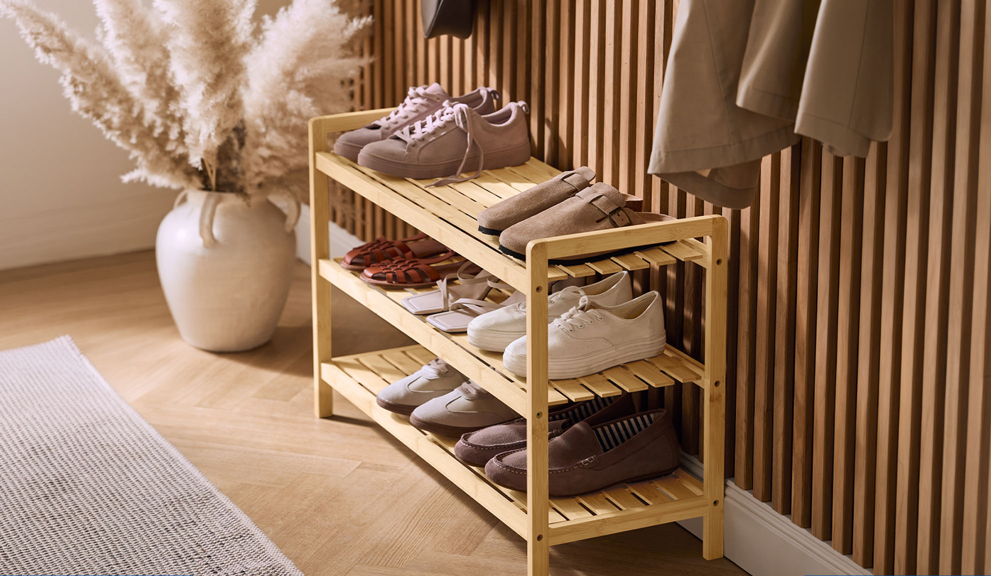Wooden shoe rack with various shoes and a pampas grass vase in a hallway.