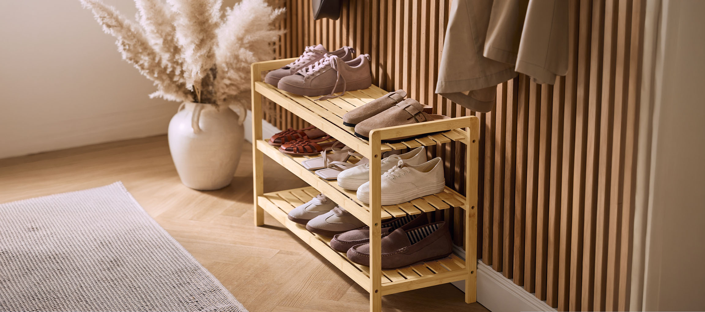 Wooden shoe rack with various shoes, a pampas grass vase, and a coat on the wall.