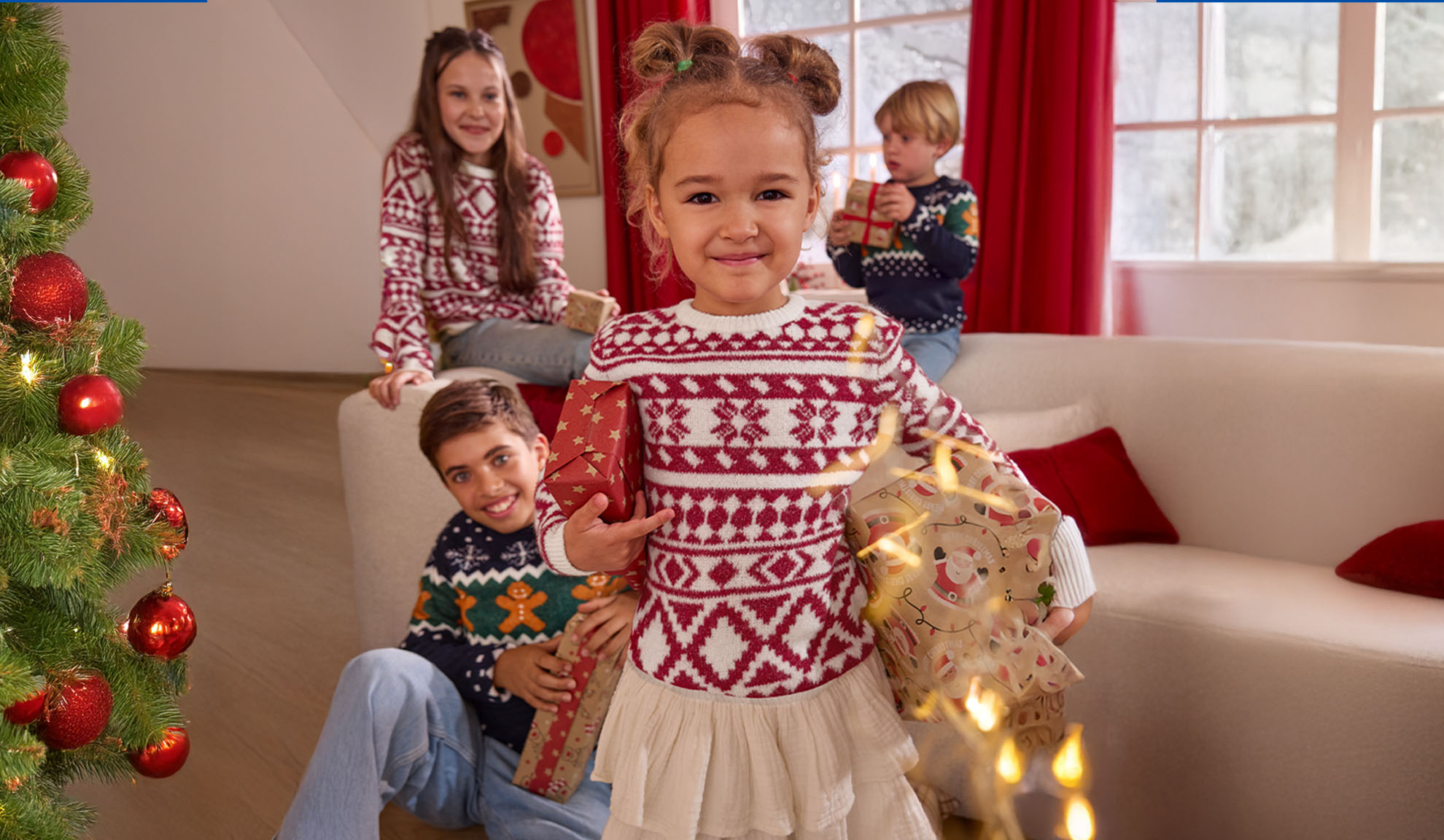 Four children in Christmas sweaters holding gifts, with a decorated Christmas tree.