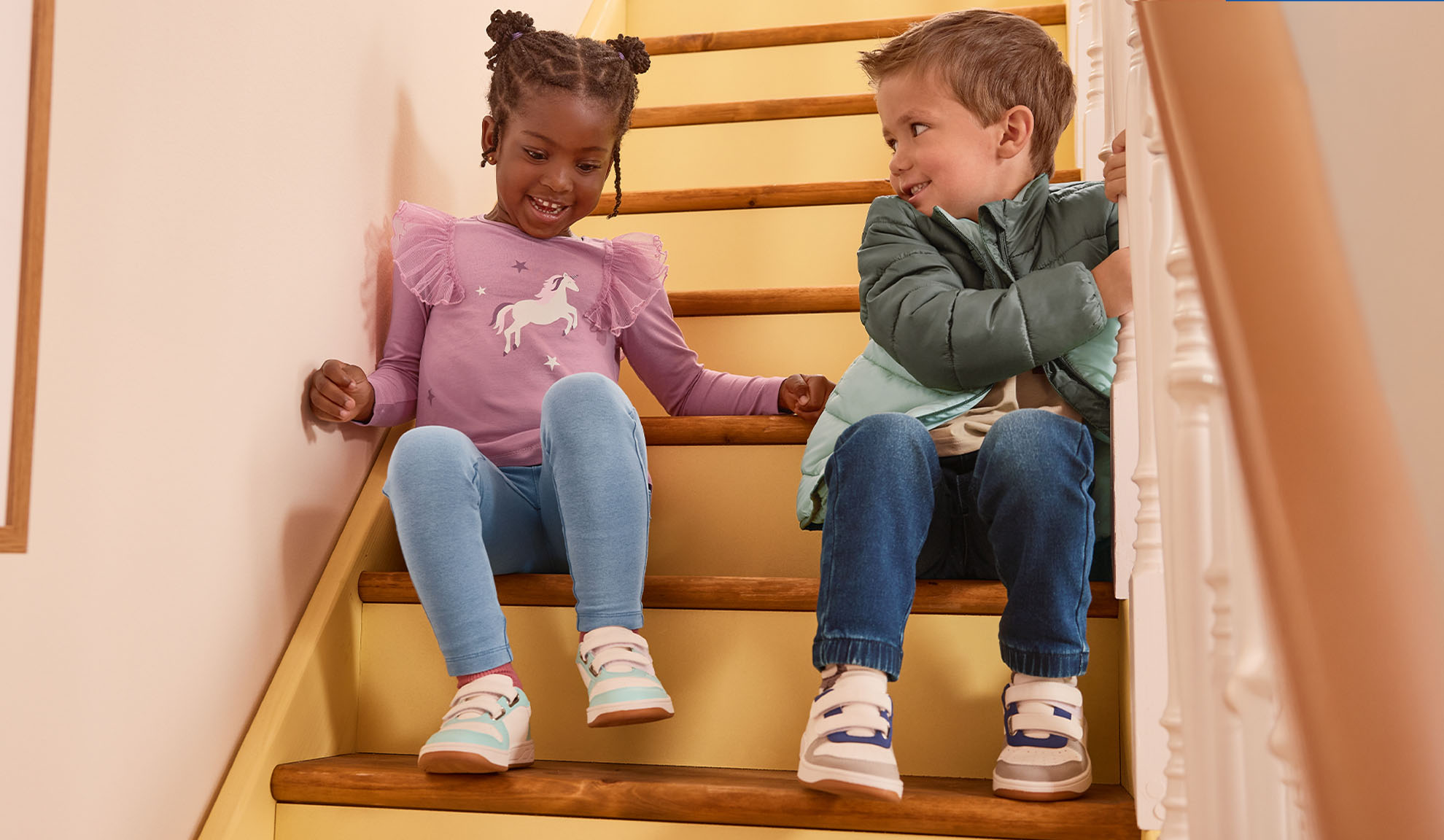 Two children in casual clothes and sneakers sitting on wooden stairs, smiling at each other.