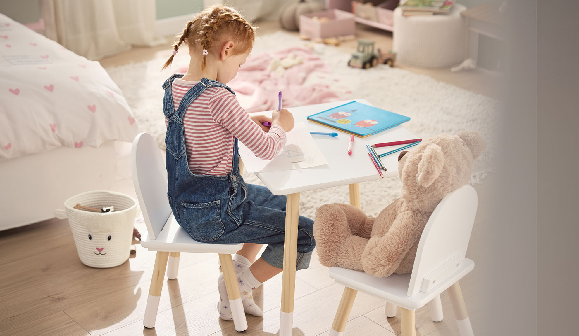 Girl drawing at a white table with a teddy bear, in a child's room.