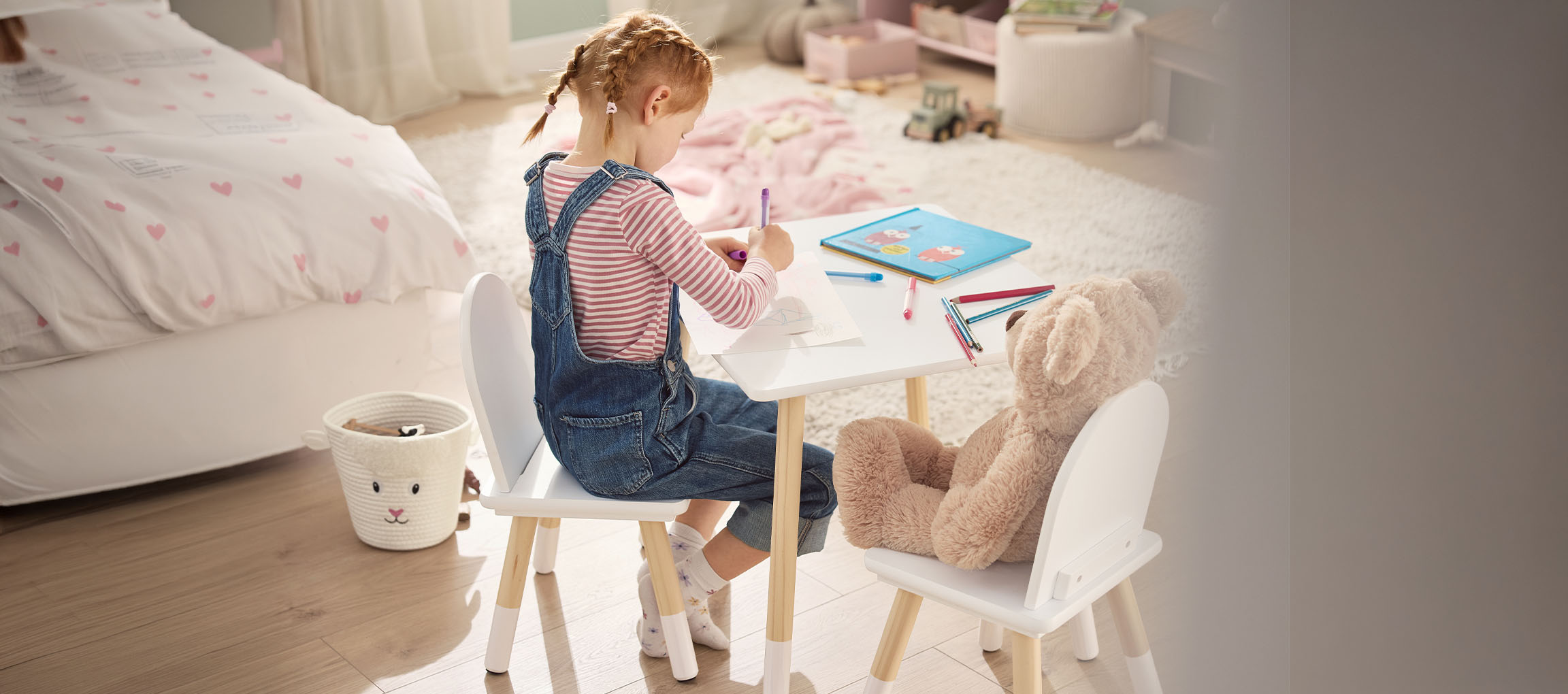 Girl drawing at a white table with chairs, a teddy bear next to her.