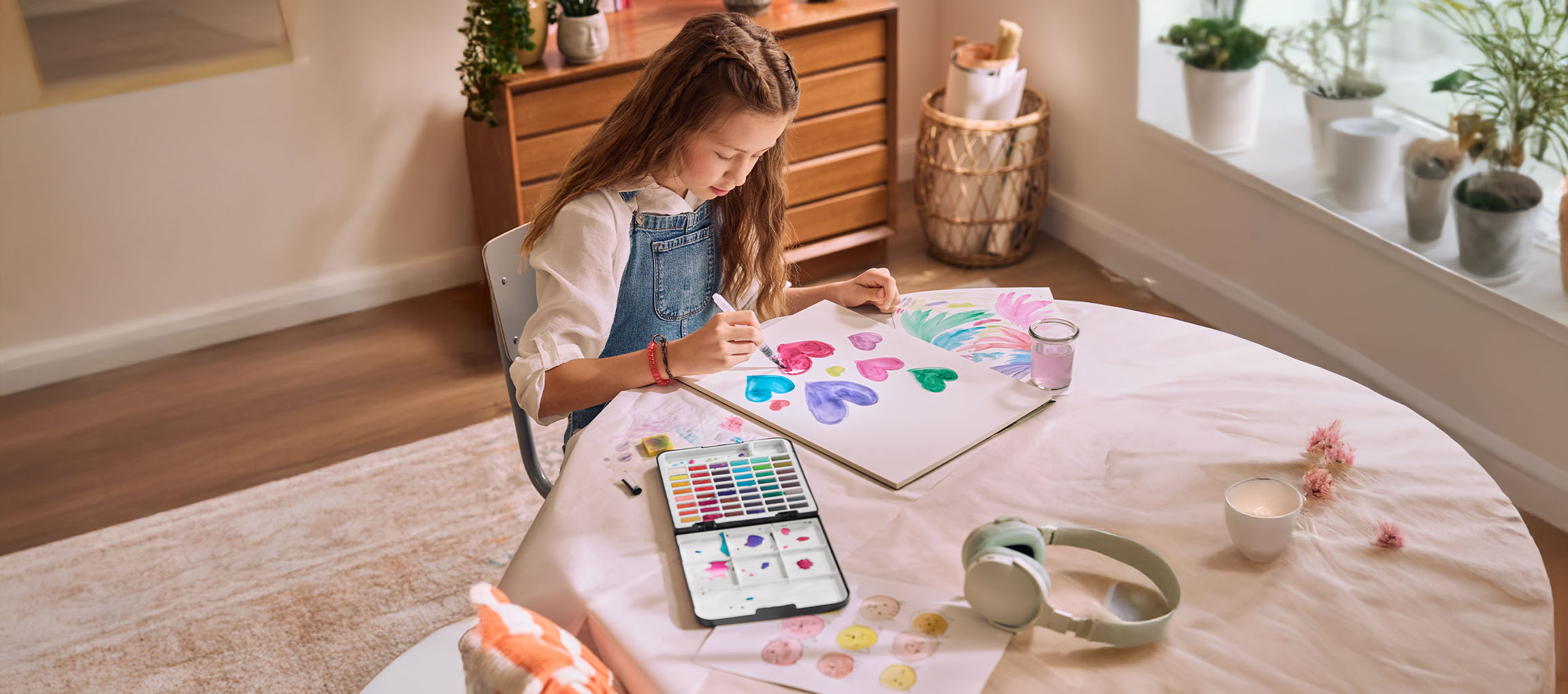 A girl painting hearts with watercolors at a table, with a tablet displaying a color palette.