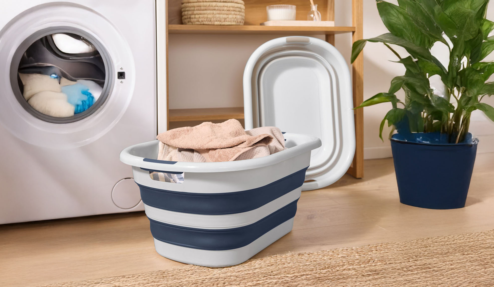 A washing machine with clothes, a collapsible laundry basket, and a plant.
