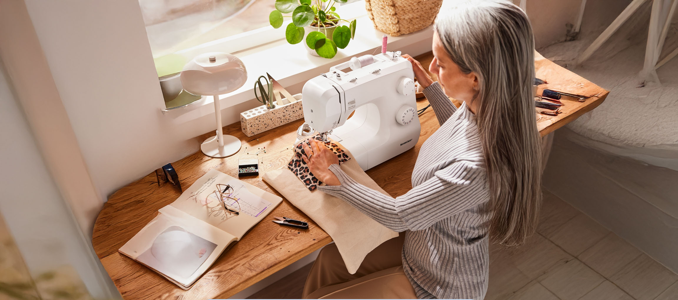 A woman sews on a SilverCrest sewing machine, with a sketch book and sewing accessories on the table.
