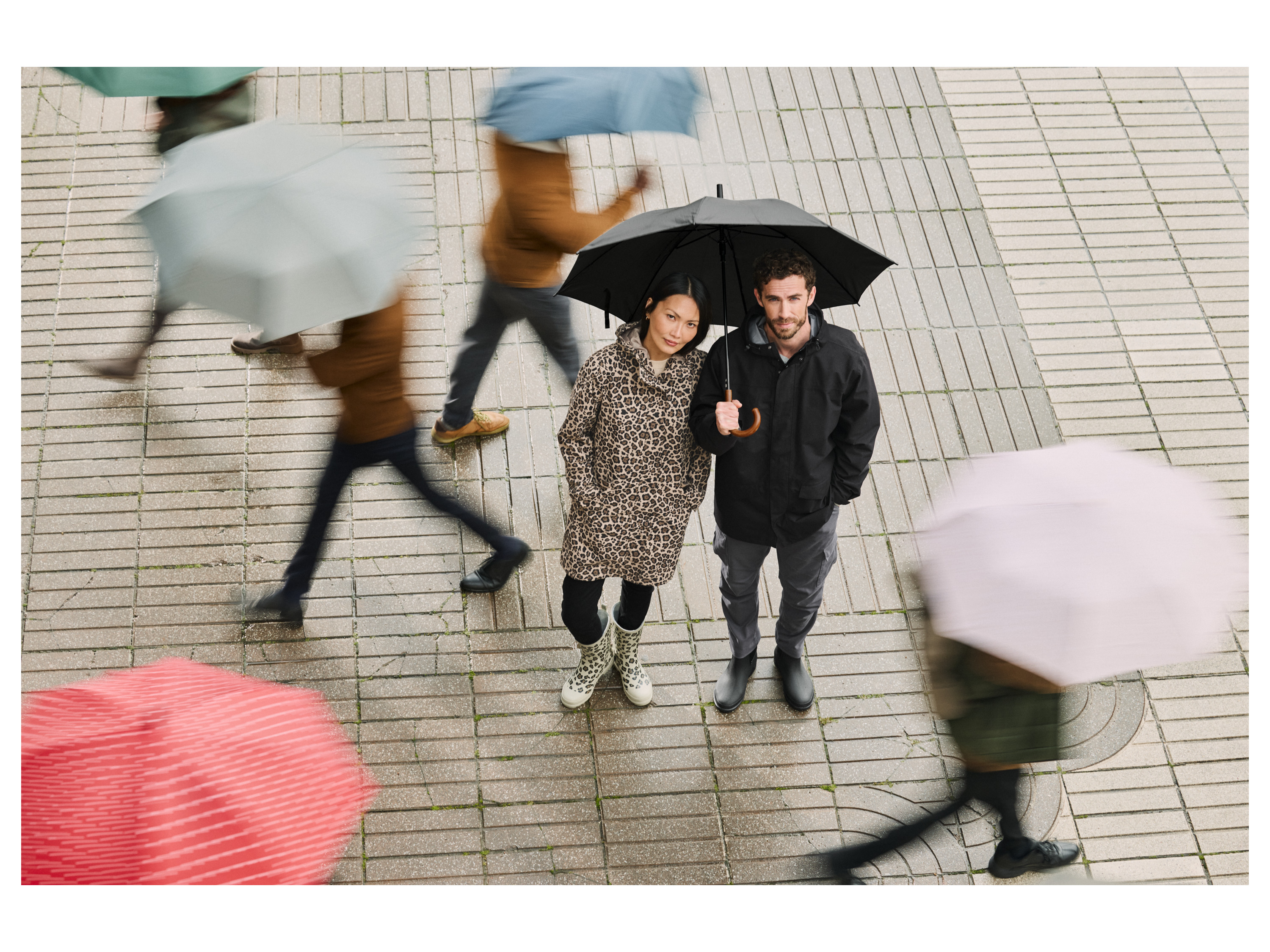 A couple in rain gear with an umbrella, surrounded by blurred figures with umbrellas.