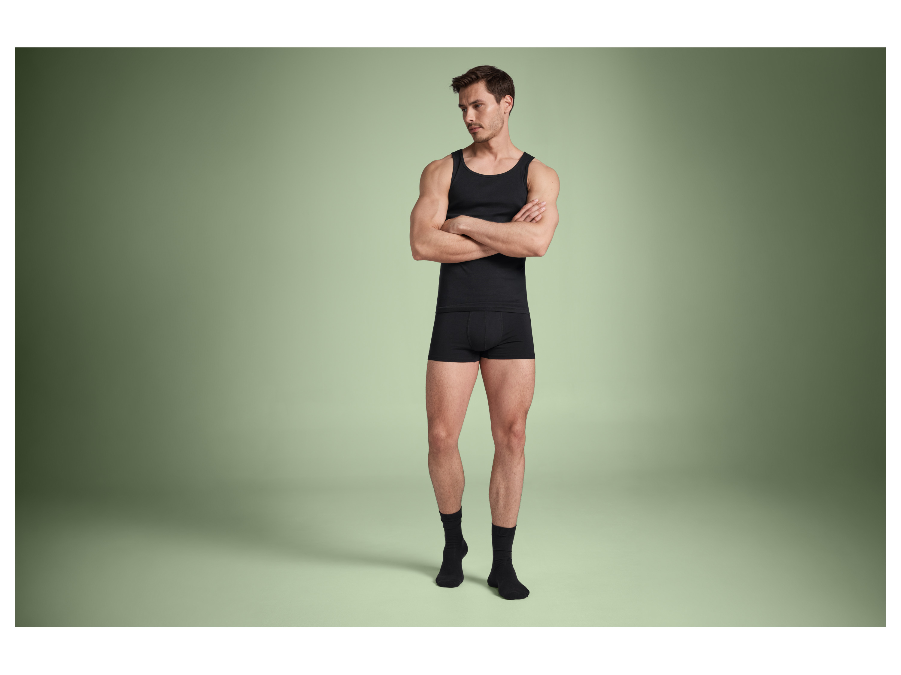 A man models black undershirt and boxer briefs against a sage green background.