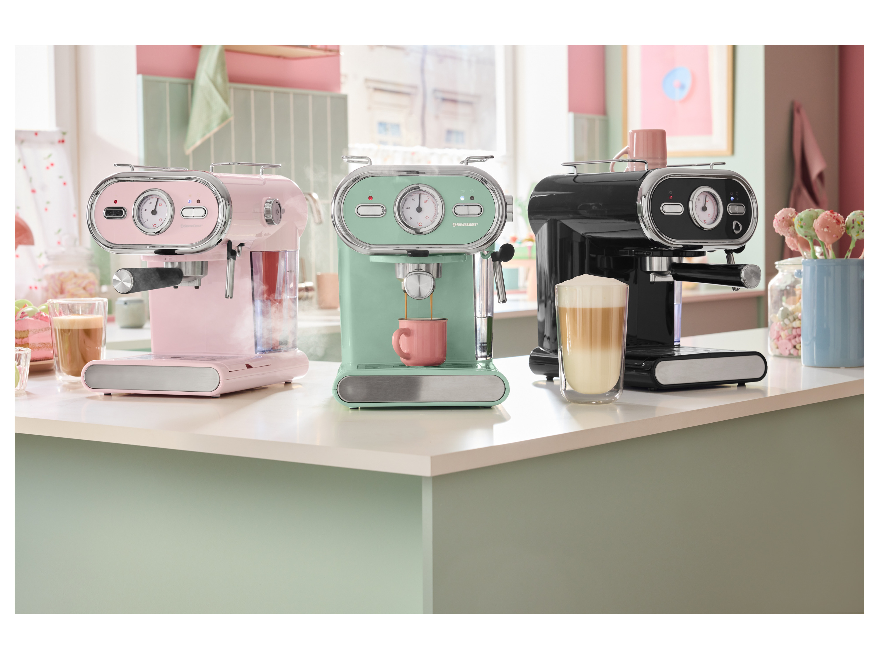 Three SilverCrest espresso machines in pink, mint, and black on a kitchen counter.
