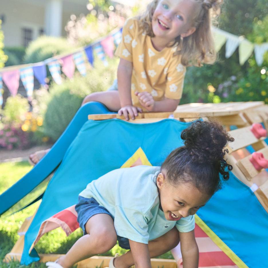 Two happy children playing on a wooden climbing frame with a blue tent in a sunny garden.