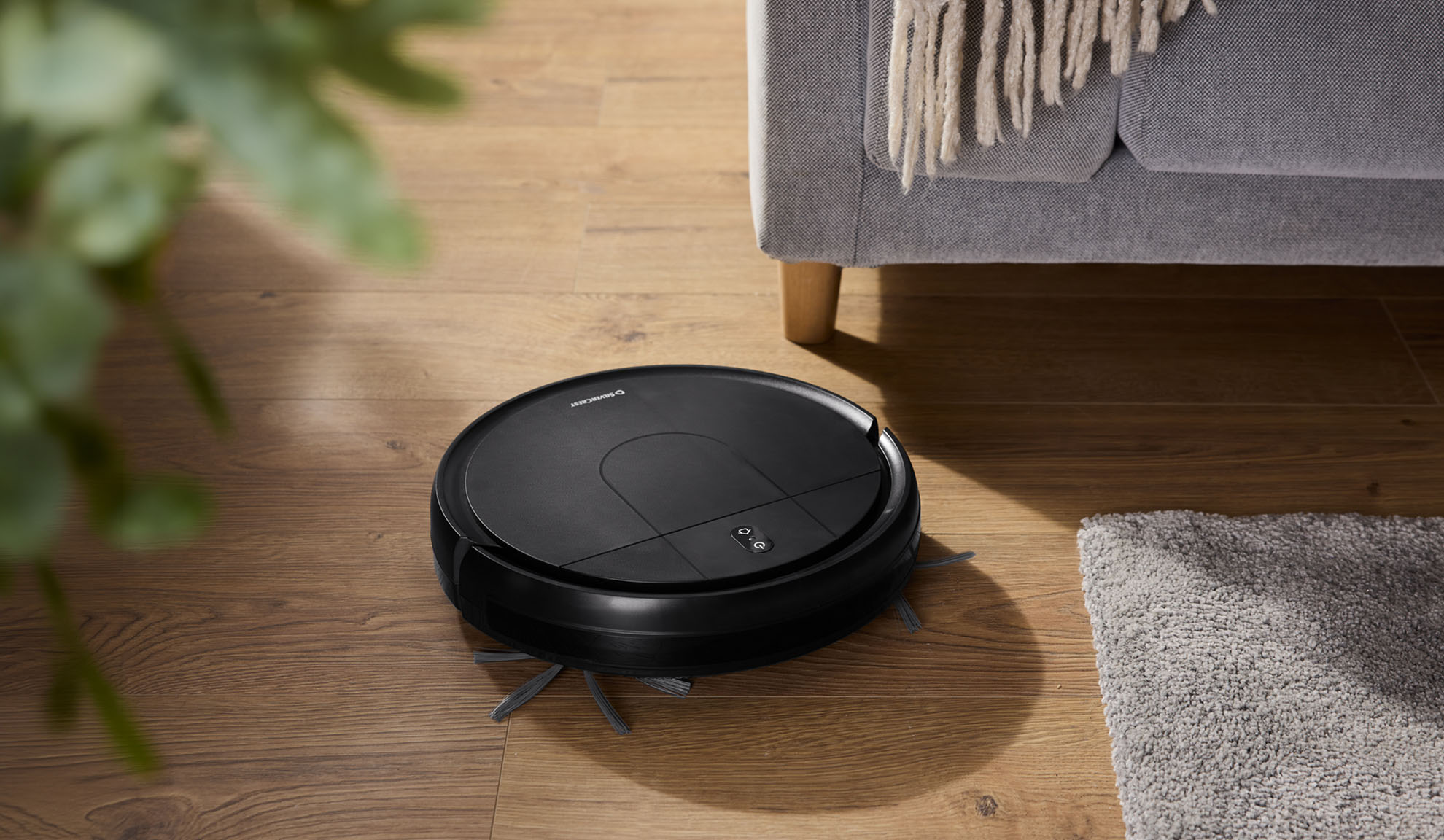Black robotic vacuum cleaner on a wooden floor next to a grey sofa and rug.