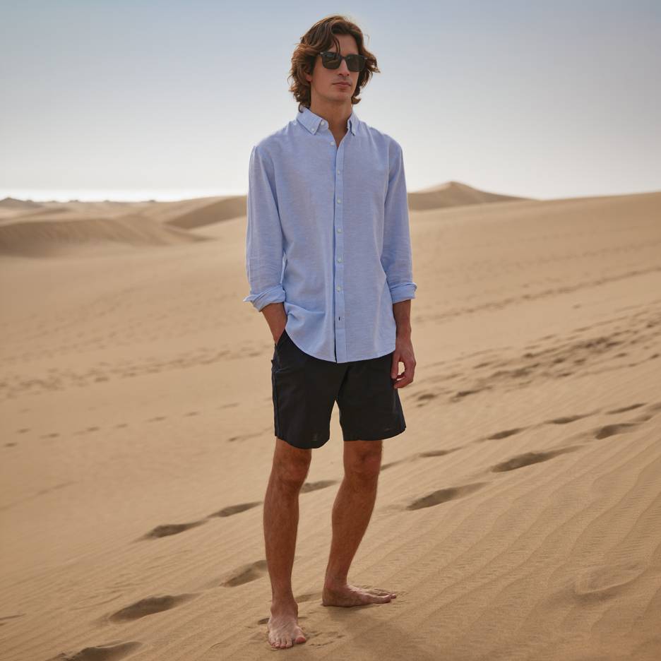 Man in blue shirt and black shorts, barefoot standing on sand dunes.