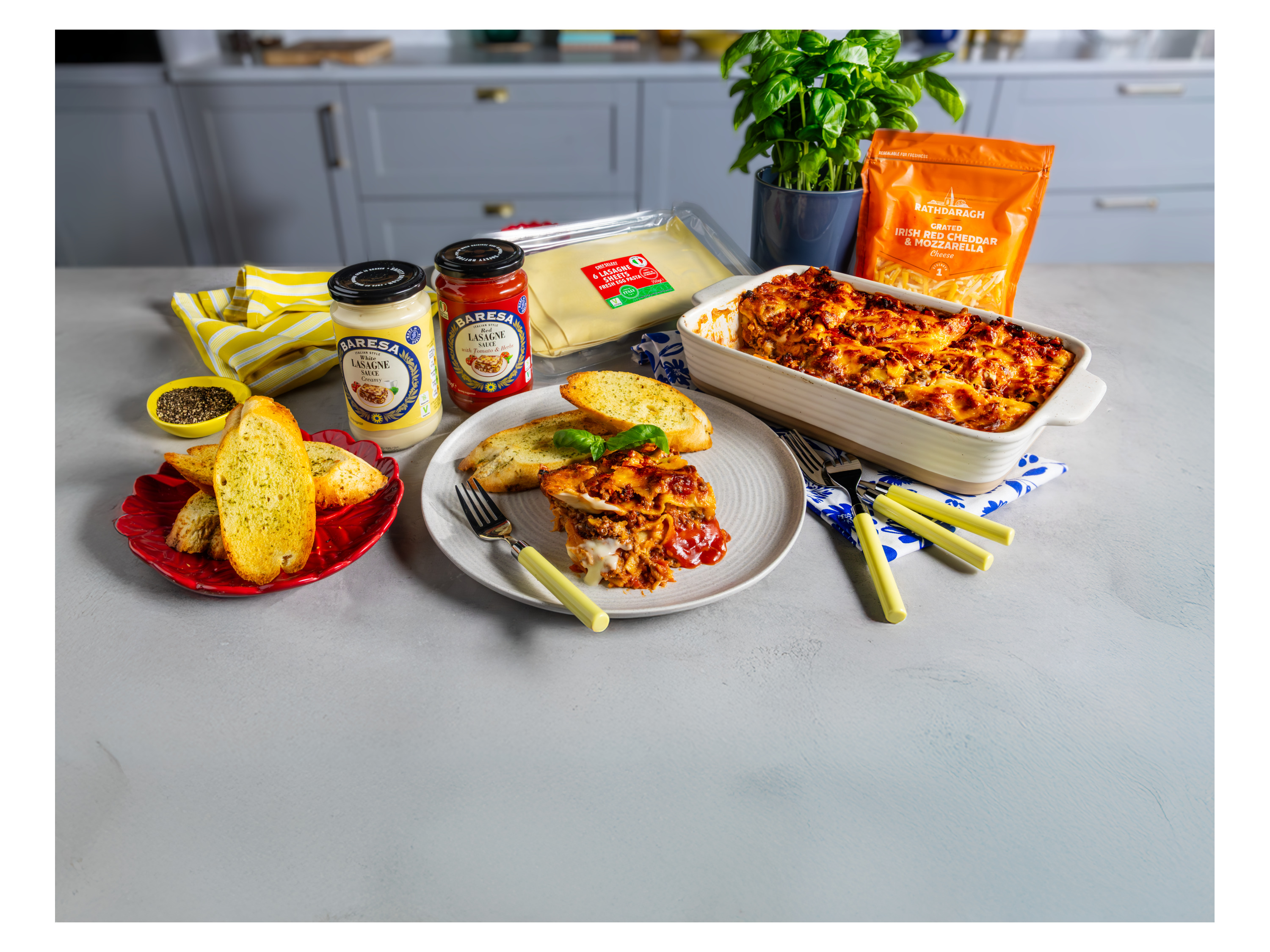 Lasagna dish with ingredients like sauces, pasta sheets, cheese, and garlic bread on a kitchen counter.
