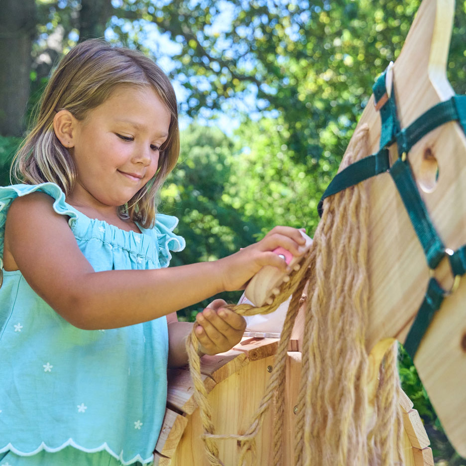 Girl brushing the mane of a wooden rocking horse outdoors.