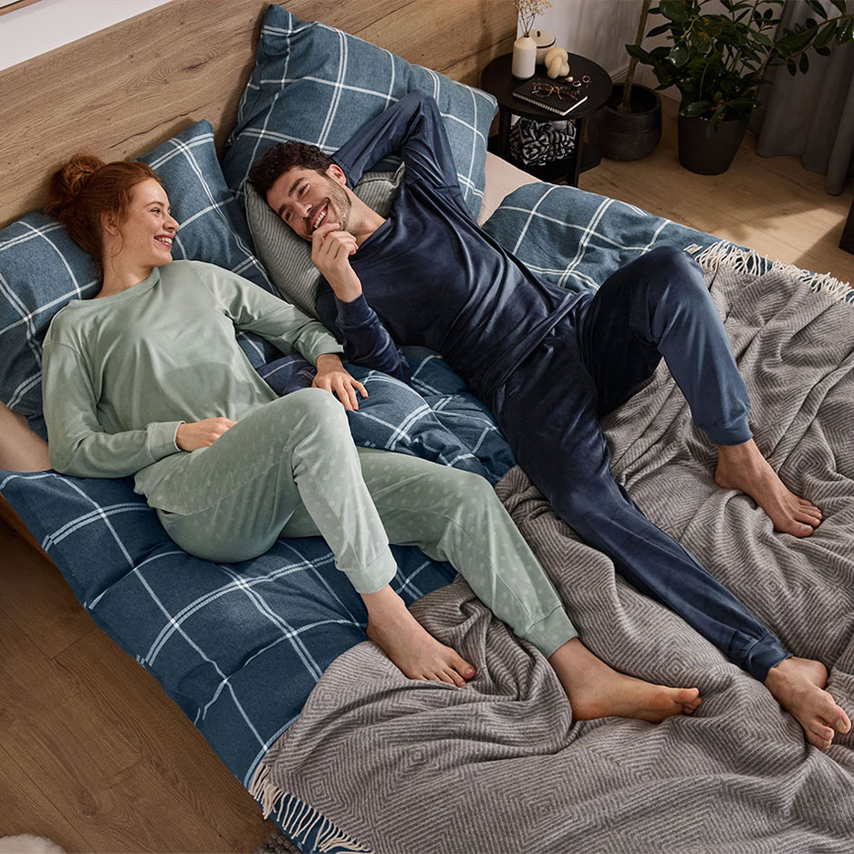 Couple in pajamas lying on a bed with plaid bedding and a blanket.