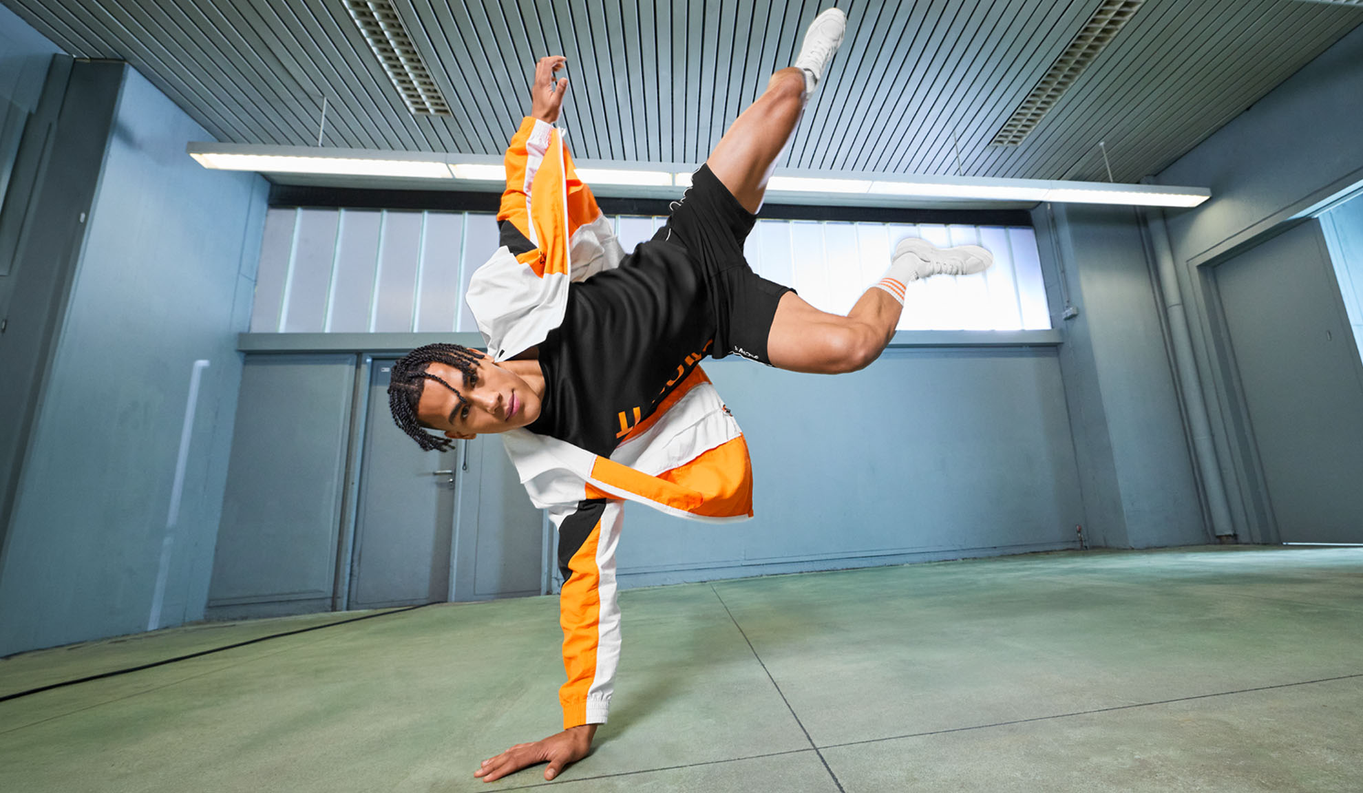 Man in sportswear, black t-shirt, shorts, and orange and white jacket, performing a dance move.