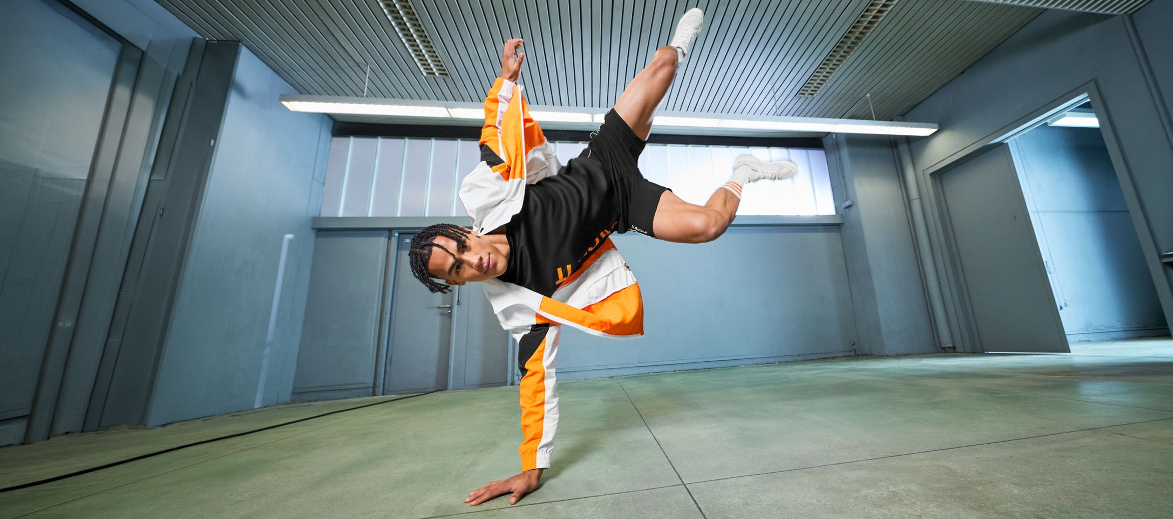 Man in sportswear, black t-shirt and orange and white jacket, dancing in a room.
