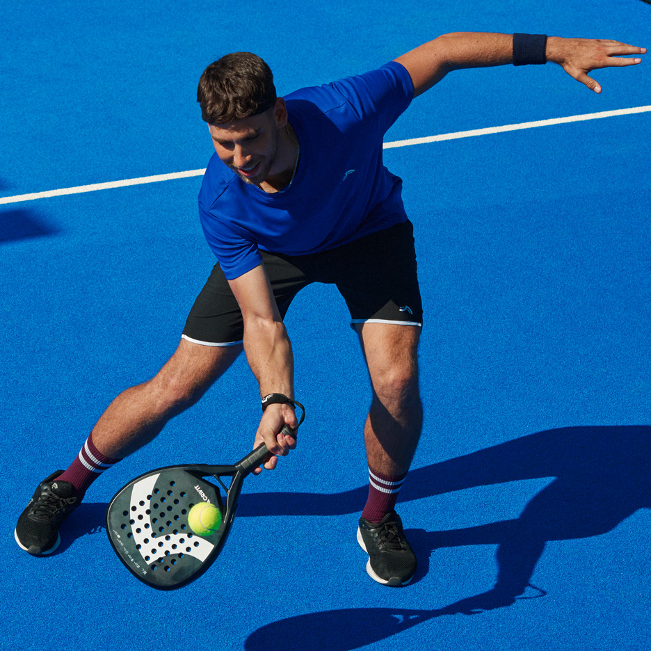 Man in blue t-shirt and black shorts playing padel with a racket and ball.
