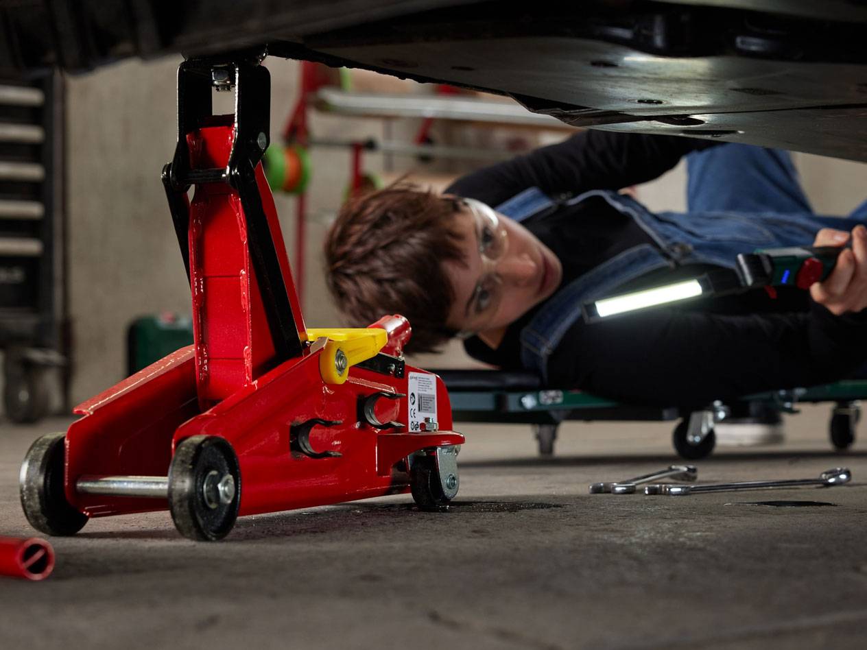 Red hydraulic jack and woman under a car with a work light and tools.