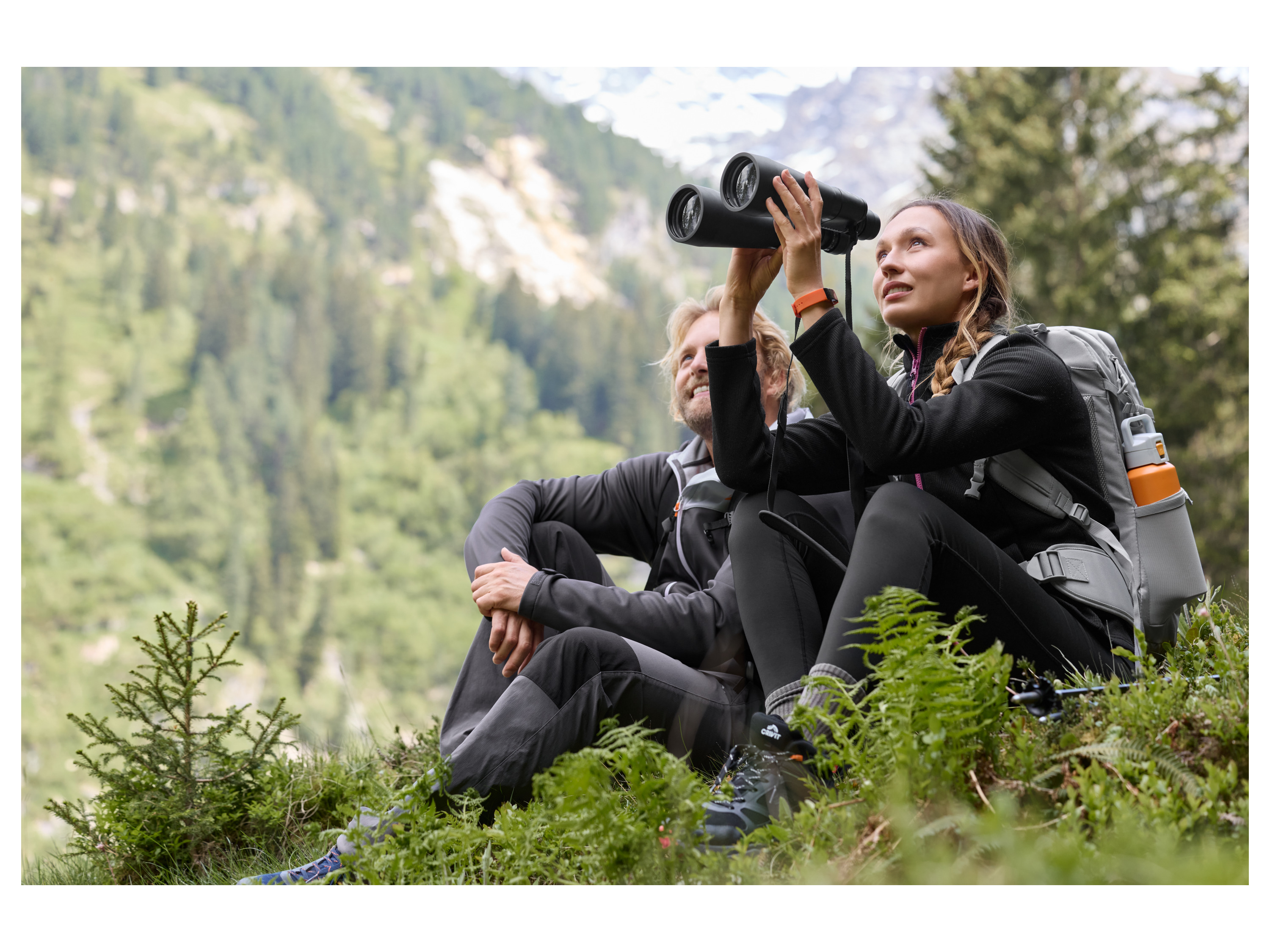 Two hikers with binoculars and backpacks enjoying a mountain view.