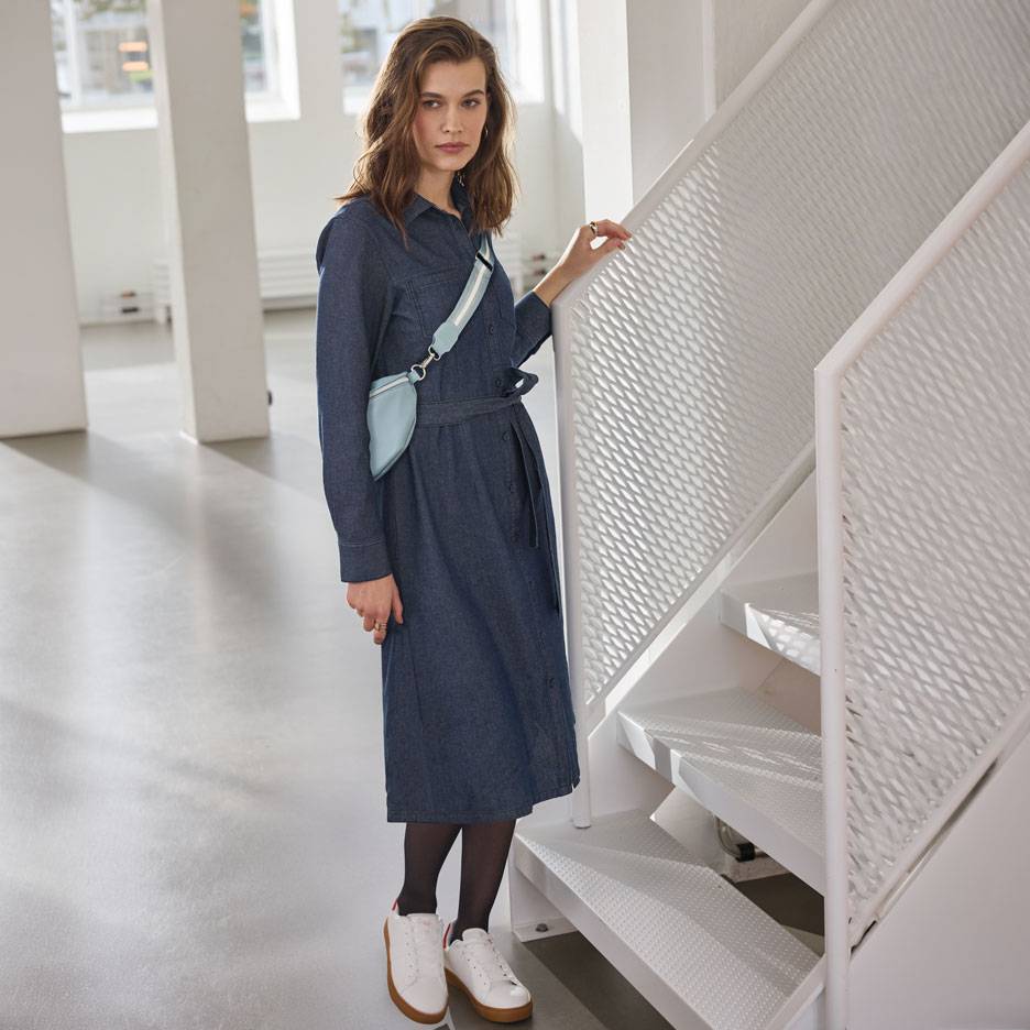 Woman in a denim shirt dress, black tights, and white sneakers, with a light blue fanny pack.